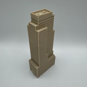 May include: A beige, multi-story skyscraper model with a rectangular base and a stepped design. The building features numerous small, square windows and a flat roof. The model is set against a plain, light gray background.