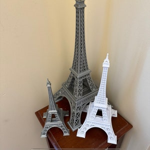 May include: Three Eiffel Tower models, one large gray model and two smaller white models, on a wooden table with a ruler.