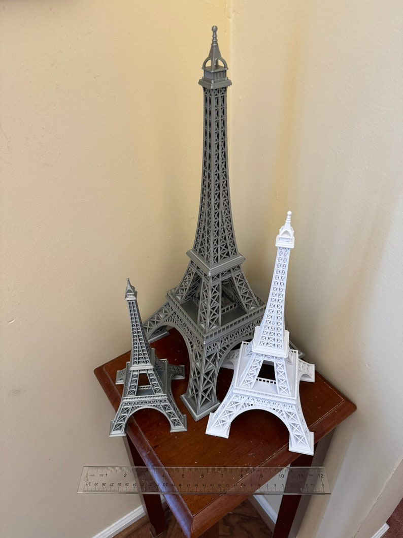May include: Three Eiffel Tower models, one large gray model and two smaller white models, on a wooden table with a ruler.