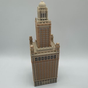 May include: A miniature architectural model of a skyscraper featuring a tan and brown facade with numerous vertical window columns, a tiered rectangular base, four corner turrets, and a central tower topped with a rounded dome structure.