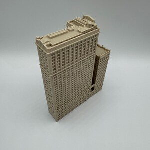 May include: A beige architectural model of a tall building with many windows. The building has a rectangular shape with a flat roof and detailed facade. The model is a miniature replica of a skyscraper.