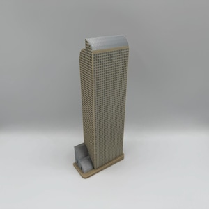 May include: A tan and gray miniature skyscraper model. The building has a rectangular shape with a grid-like pattern on the facade. The top of the building is a lighter gray color. The model includes a smaller rectangular base.