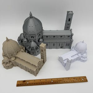 May include: Four miniature architectural models of the Florence Cathedral, including the dome and bell tower. The models are in shades of gray, beige, and white. A wooden ruler, approximately 12 inches long, is placed near the models.