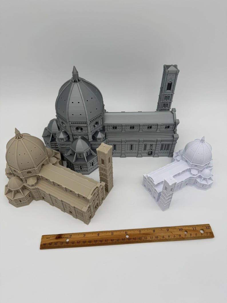 May include: Four miniature architectural models of the Florence Cathedral, including the dome and bell tower. The models are in shades of gray, beige, and white. A wooden ruler, approximately 12 inches long, is placed near the models.