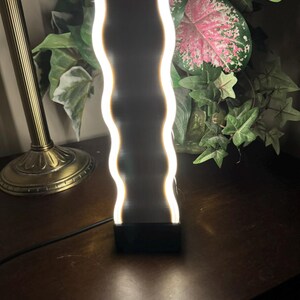 K&ouml;nnte beinhalten: Eine moderne, wellenf&ouml;rmige LED-Lampe mit schwarzem Sockel und wei&szlig;em, leuchtendem Licht. Die Lampe steht auf einer dunklen Holzoberfl&auml;che, mit einer goldfarbenen Lampe und Gr&uuml;nzeug im Hintergrund. Die Lampe ist etwa 25 cm hoch.