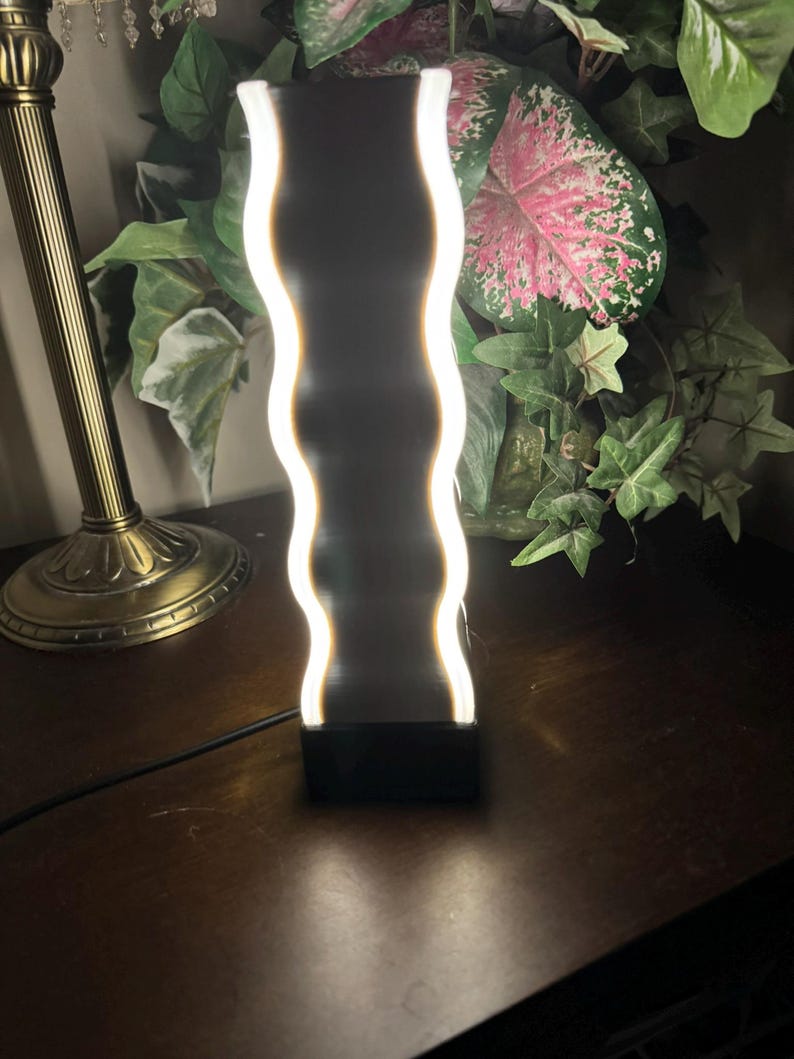 K&ouml;nnte beinhalten: Eine moderne, wellenf&ouml;rmige LED-Lampe mit schwarzem Sockel und wei&szlig;em, leuchtendem Licht. Die Lampe steht auf einer dunklen Holzoberfl&auml;che, mit einer goldfarbenen Lampe und Gr&uuml;nzeug im Hintergrund. Die Lampe ist etwa 25 cm hoch.
