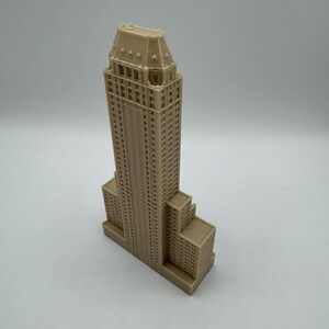 Może przedstawiać: Beżowy miniaturowy model wieżowca ze szczeg&oacute;łową fasadą. Budynek ma prostokątną podstawę z wieloma poziomami i wysoką, ozdobną wieżą. Model wykonany jest z litego materiału i stoi na białej powierzchni.