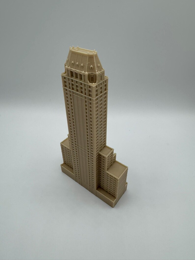 Może przedstawiać: Beżowy miniaturowy model wieżowca ze szczeg&oacute;łową fasadą. Budynek ma prostokątną podstawę z wieloma poziomami i wysoką, ozdobną wieżą. Model wykonany jest z litego materiału i stoi na białej powierzchni.