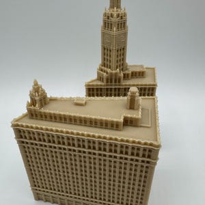 May include: A tan-colored architectural model of a building with a tall clock tower. The model has detailed features, including windows and decorative elements. The building's design is reminiscent of a historic skyscraper.