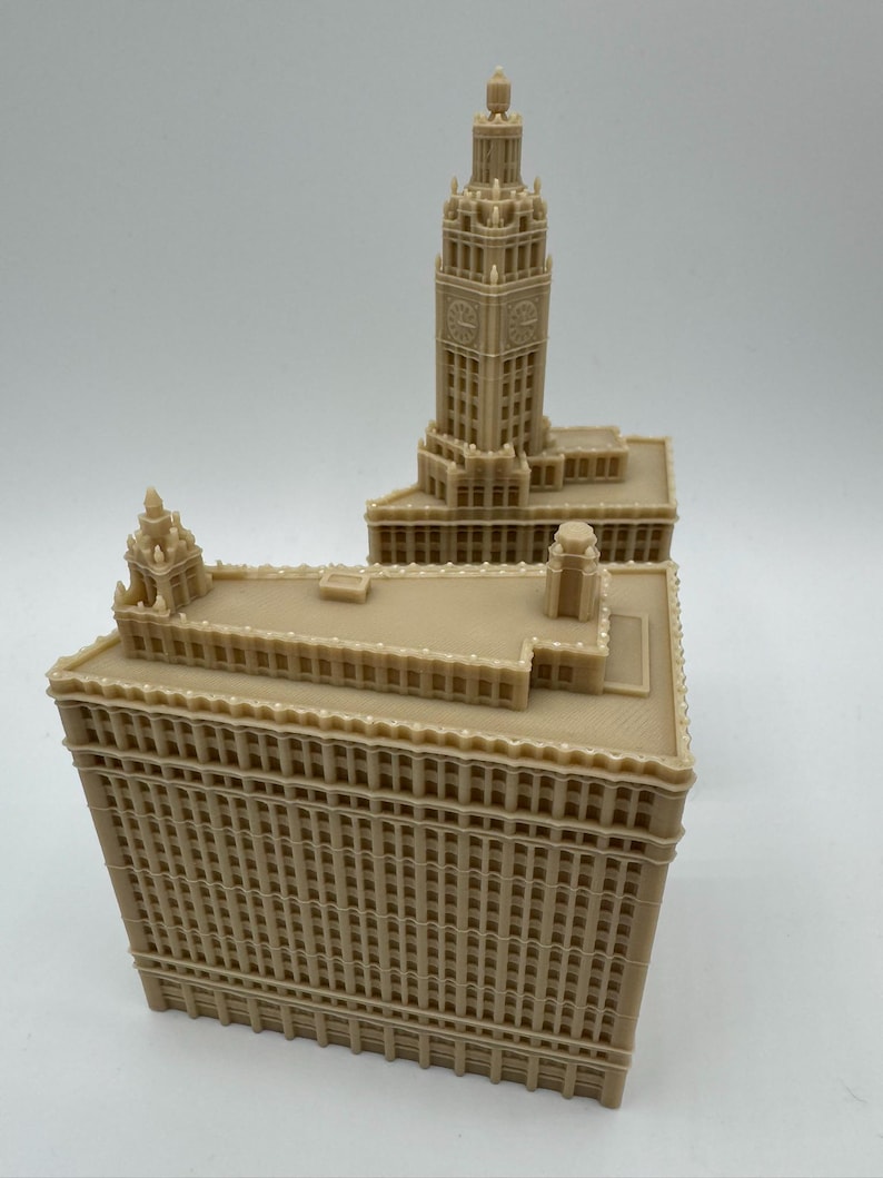 May include: A tan-colored architectural model of a building with a tall clock tower. The model has detailed features, including windows and decorative elements. The building's design is reminiscent of a historic skyscraper.