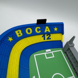 May include: A 3D-printed model of a football stadium, with a green pitch and white lines. The stands are blue and yellow, with the word "BOCA" in yellow and the number "12". A grey tower is visible.