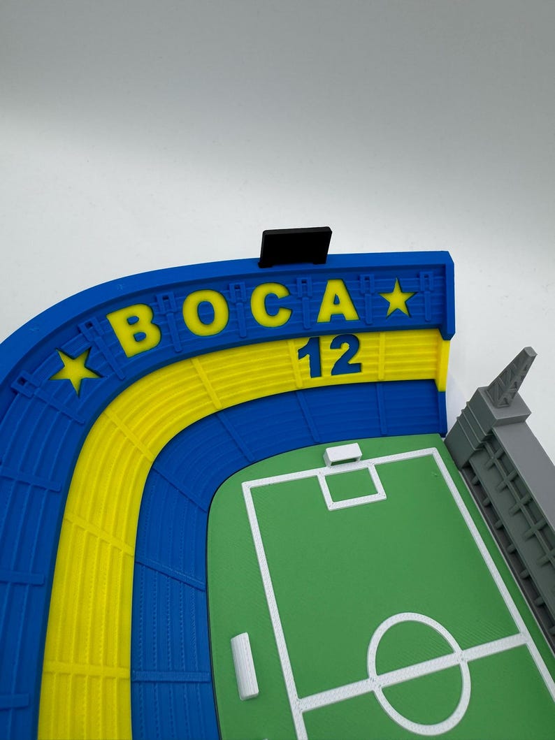 May include: A 3D-printed model of a football stadium, with a green pitch and white lines. The stands are blue and yellow, with the word "BOCA" in yellow and the number "12". A grey tower is visible.