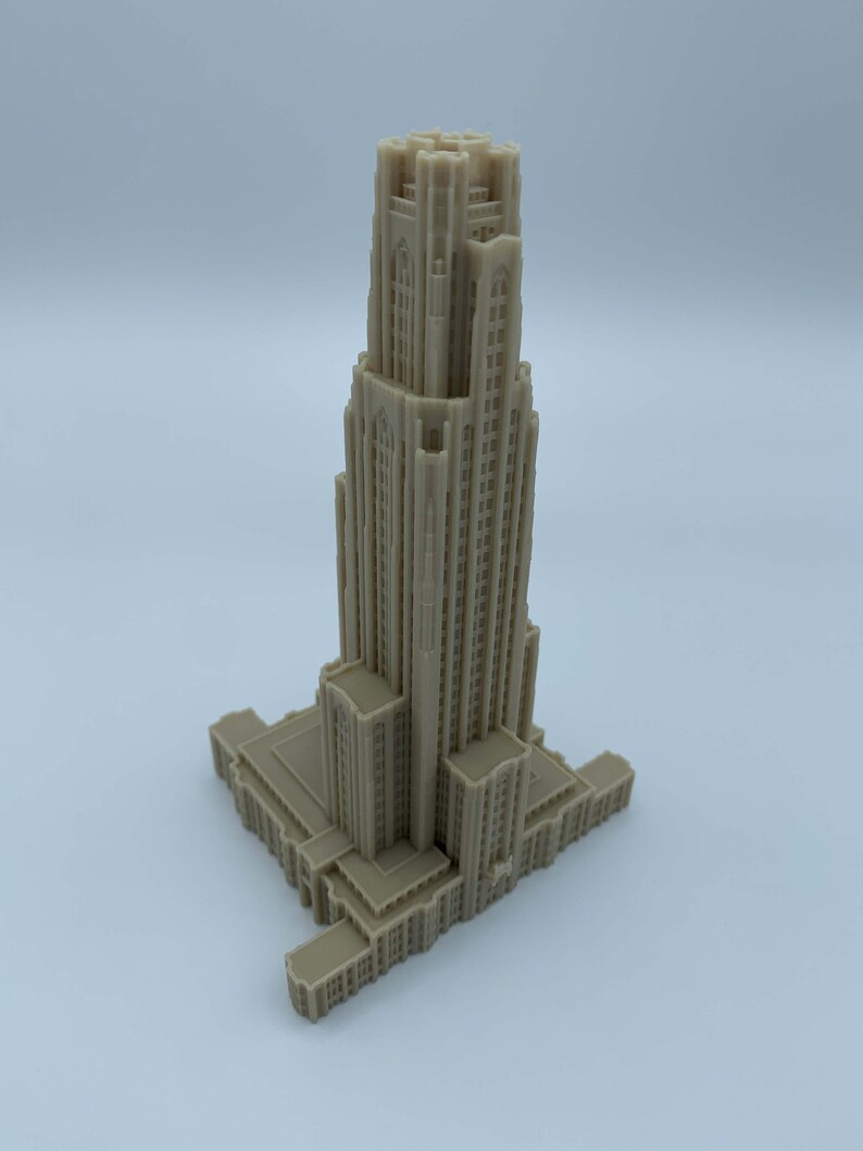 May include: A detailed 3D printed model of a tall, beige building with a pointed roof and many windows. The building has a rectangular base and is set against a plain white background.
