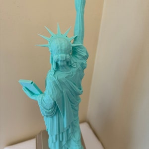 May include: A teal blue 3D printed model of the Statue of Liberty. The model is standing on a gray base.