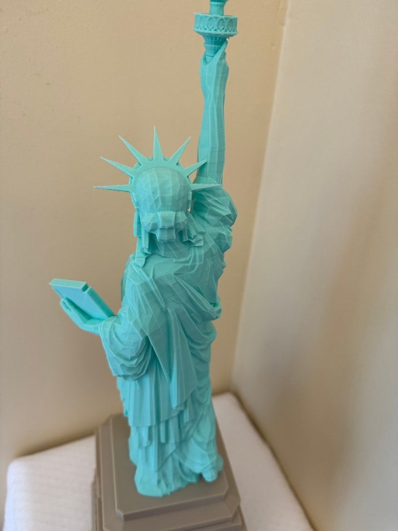 May include: A teal blue 3D printed model of the Statue of Liberty. The model is standing on a gray base.