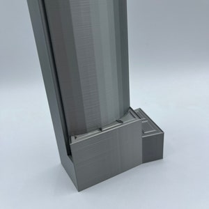 May include: A gray 3D printed object with a rectangular base and a tall, cylindrical tower. The base has a series of steps or ledges.