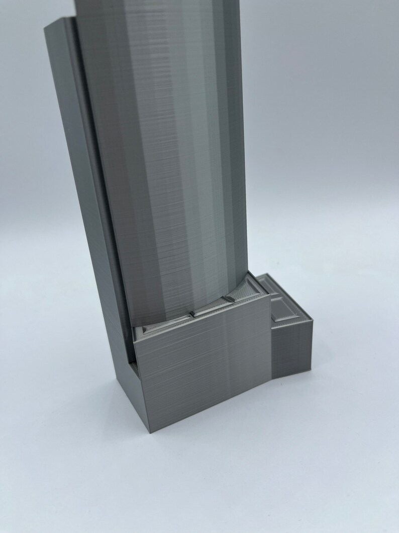 May include: A gray 3D printed object with a rectangular base and a tall, cylindrical tower. The base has a series of steps or ledges.