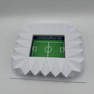 May include: A white, geometric stadium model with a green soccer field. The stadium has a detailed, angular design and the text "TRABIZUNISPOIR" is visible. The field features white markings and a central circle.