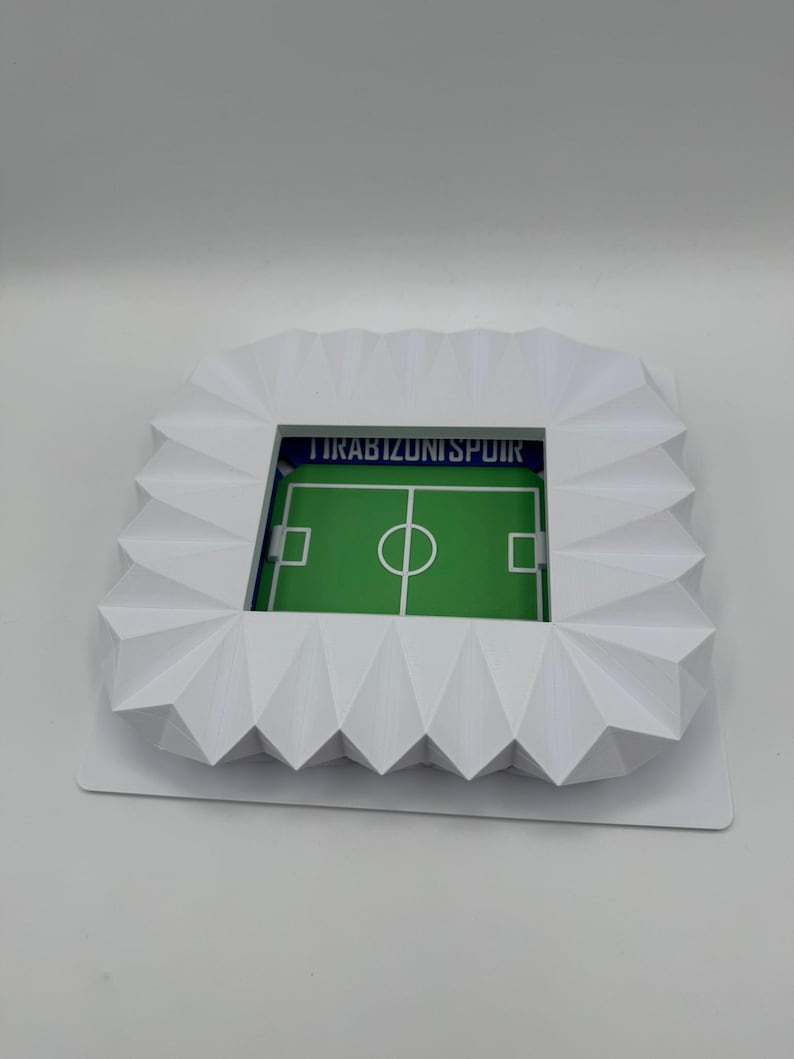 May include: A white, geometric stadium model with a green soccer field. The stadium has a detailed, angular design and the text "TRABIZUNISPOIR" is visible. The field features white markings and a central circle.