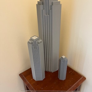 May include: Three gray 3D printed models of skyscrapers, the tallest is about 12 inches tall, the middle is about 8 inches tall, and the smallest is about 5 inches tall. The models are sitting on a wooden table.