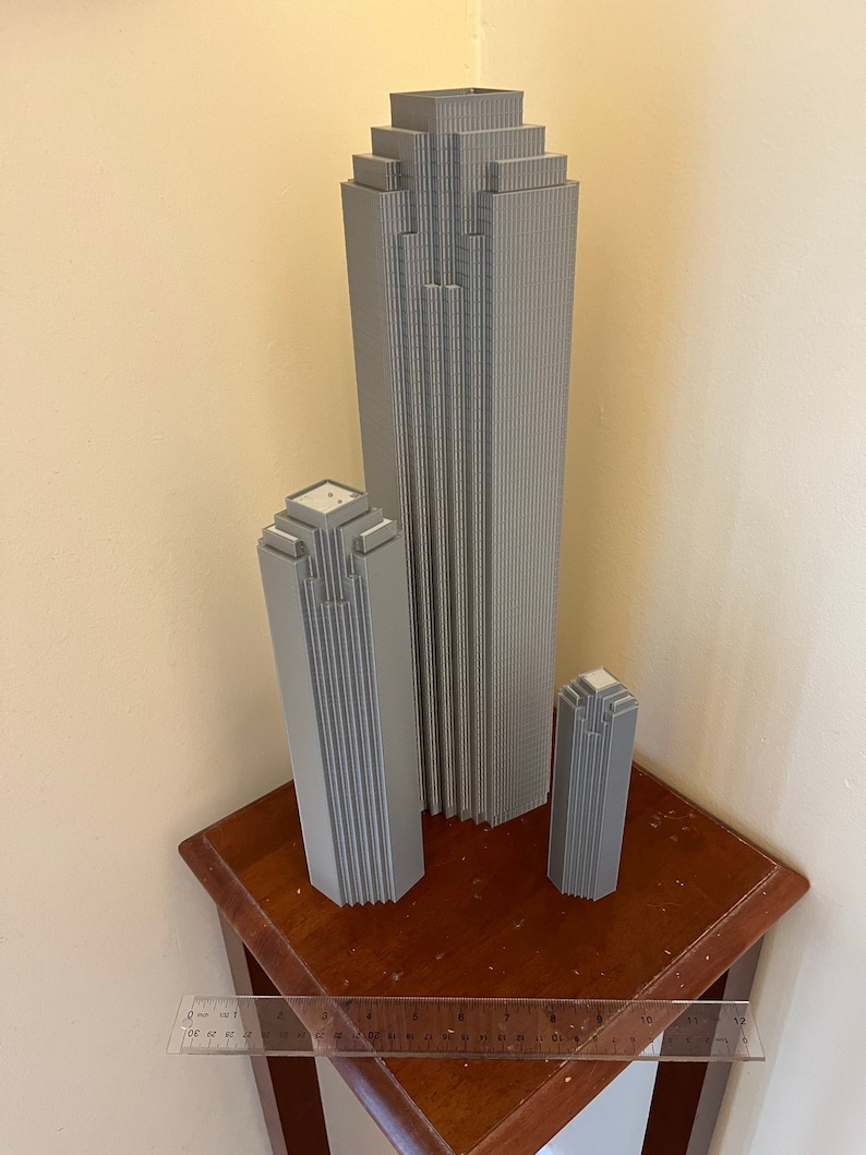 May include: Three gray 3D printed models of skyscrapers, the tallest is about 12 inches tall, the middle is about 8 inches tall, and the smallest is about 5 inches tall. The models are sitting on a wooden table.