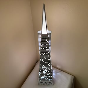 May include: A silver-coloured, illuminated skyscraper model with a pointed top. The building's windows are represented by small, bright white squares. The model is placed on a white surface, with a beige wall in the background.