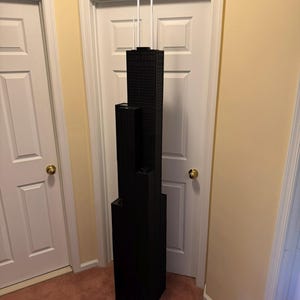 May include: A black architectural model of a skyscraper, featuring multiple rectangular sections stacked vertically. The model is suspended from the ceiling by white cords, showcasing a detailed facade. The model is approximately 122 cm tall.