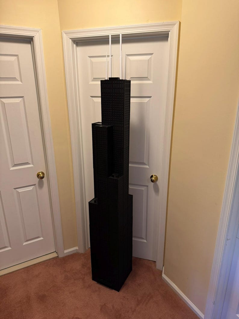 May include: A black architectural model of a skyscraper, featuring multiple rectangular sections stacked vertically. The model is suspended from the ceiling by white cords, showcasing a detailed facade. The model is approximately 122 cm tall.