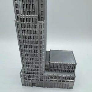 May include: A gray, 3D-printed model of a skyscraper. The building features many windows and architectural details, with a rectangular base and a tall, multi-story tower. The model is set against a plain white background.