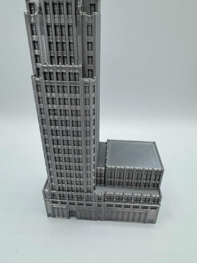 May include: A gray, 3D-printed model of a skyscraper. The building features many windows and architectural details, with a rectangular base and a tall, multi-story tower. The model is set against a plain white background.