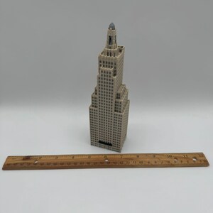 May include: A beige miniature skyscraper model with a dark top. The building has many windows and is placed on a white surface. A wooden ruler is placed in front of the building.