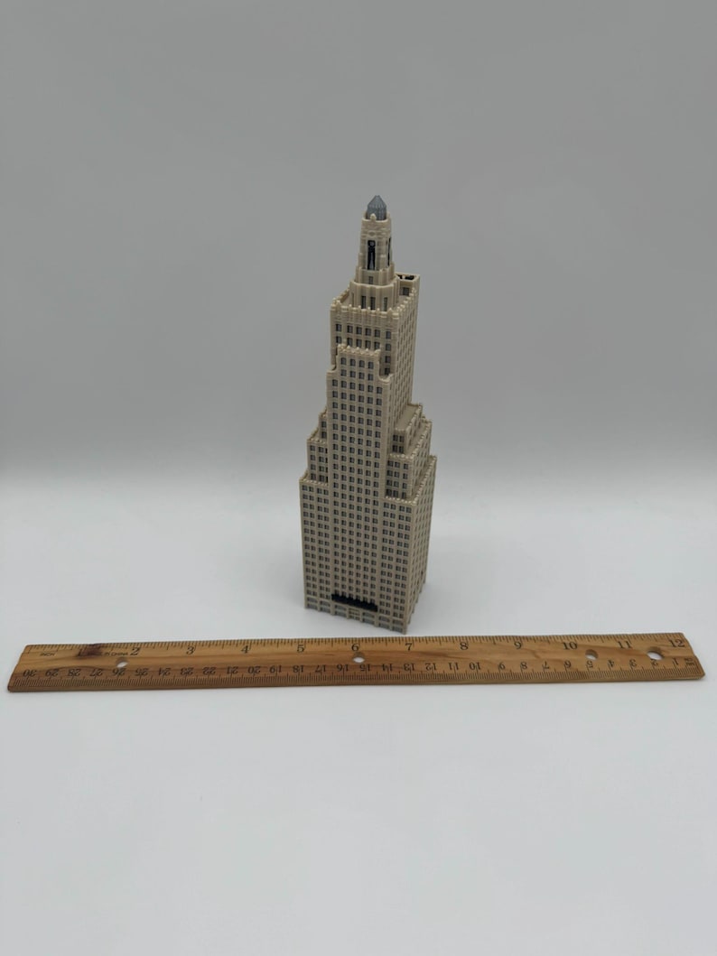 May include: A beige miniature skyscraper model with a dark top. The building has many windows and is placed on a white surface. A wooden ruler is placed in front of the building.