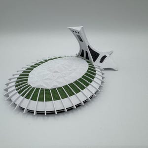 Puede incluir: Maqueta arquitect&oacute;nica blanca y verde con un dise&ntilde;o &uacute;nico. El modelo presenta una base circular con una parte superior texturizada y aletas blancas. Una estructura de soporte blanca con detalles negros est&aacute; unida a la base.