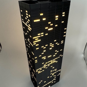 Peut inclure: Une lampe noire en forme de gratte-ciel, avec des fen&ecirc;tres rectangulaires &eacute;clair&eacute;es. Les fen&ecirc;tres &eacute;mettent une lumi&egrave;re jaune chaude, cr&eacute;ant un effet de paysage urbain nocturne. La lampe a un cordon d'alimentation blanc. D&eacute;coration d'int&eacute;rieur.