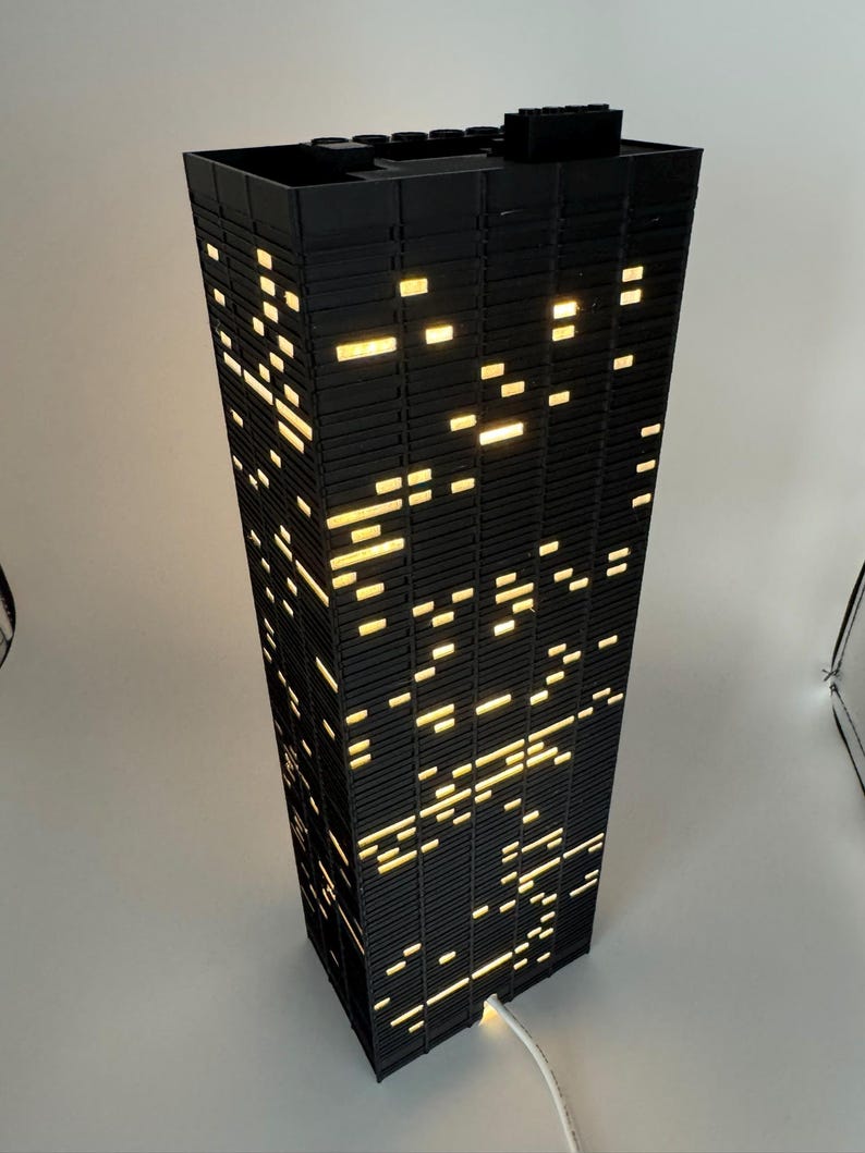 Peut inclure: Une lampe noire en forme de gratte-ciel, avec des fen&ecirc;tres rectangulaires &eacute;clair&eacute;es. Les fen&ecirc;tres &eacute;mettent une lumi&egrave;re jaune chaude, cr&eacute;ant un effet de paysage urbain nocturne. La lampe a un cordon d'alimentation blanc. D&eacute;coration d'int&eacute;rieur.