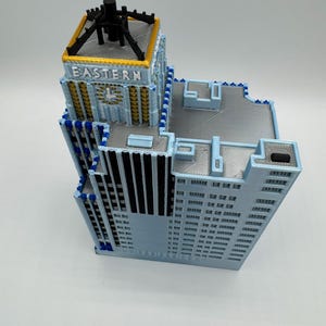 May include: A 3D-printed model of a building with a light blue facade, featuring the word "EASTERN" on the top. The model has a detailed design with multiple levels, windows, and architectural elements. The top section is black with a gold trim.