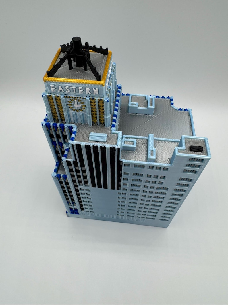 May include: A 3D-printed model of a building with a light blue facade, featuring the word "EASTERN" on the top. The model has a detailed design with multiple levels, windows, and architectural elements. The top section is black with a gold trim.
