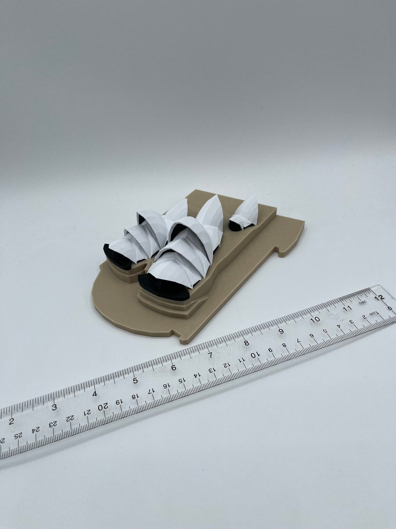 May include: A white model of the Sydney Opera House on a brown base. The model is approximately 6 inches long.