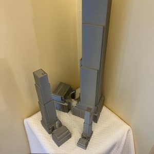May include: A 3D printed model of a skyscraper, with multiple buildings of varying heights, in a gray color. The model is on a white surface with a ruler for scale.
