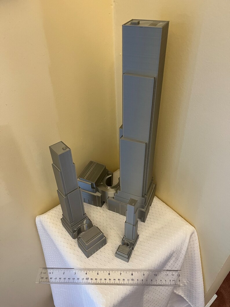 May include: A 3D printed model of a skyscraper, with multiple buildings of varying heights, in a gray color. The model is on a white surface with a ruler for scale.