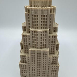 May include: A beige architectural model of a skyscraper with multiple tiers. The model has detailed window patterns and a stepped design, reminiscent of a classic Art Deco building. The structure is made of a light-coloured material.