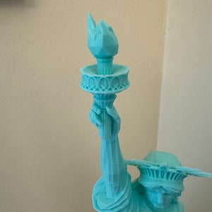 May include: A teal blue 3D printed statue of the Statue of Liberty holding a torch in her right hand.