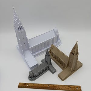 May include: Three miniature church models in white, gray, and tan are displayed. A wooden ruler is placed near the models. The models have detailed architectural features, including spires and arched windows. The ruler is approximately 12 inches long.