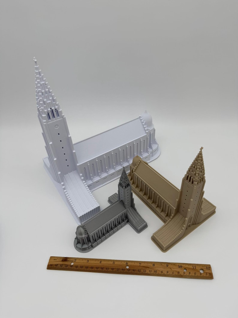 May include: Three miniature church models in white, gray, and tan are displayed. A wooden ruler is placed near the models. The models have detailed architectural features, including spires and arched windows. The ruler is approximately 12 inches long.