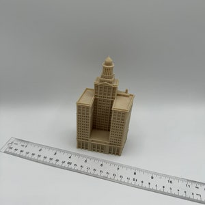 May include: A beige 3D printed model of a tall building with a dome on top. The model is about 6 inches tall and is sitting on a white surface next to a ruler.