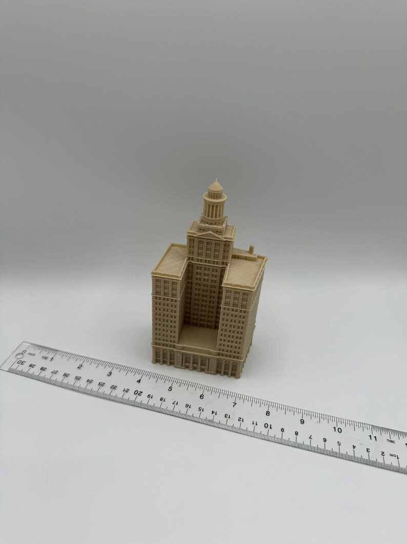 May include: A beige 3D printed model of a tall building with a dome on top. The model is about 6 inches tall and is sitting on a white surface next to a ruler.