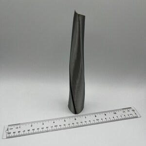 Op de afbeelding: Een zilverkleurig, gedraaid metalen beeldhouwwerk dat lijkt op een hoog gebouw. Het beeldhouwwerk is ongeveer 15 cm hoog en staat op een wit oppervlak.