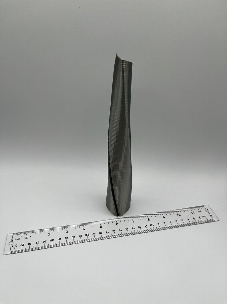 Op de afbeelding: Een zilverkleurig, gedraaid metalen beeldhouwwerk dat lijkt op een hoog gebouw. Het beeldhouwwerk is ongeveer 15 cm hoog en staat op een wit oppervlak.
