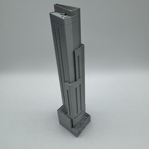 May include: A gray, miniature skyscraper model with a rectangular base and a tall, slender tower. The building has many windows and a flat roof. The model is made of a solid material and is set against a plain background.