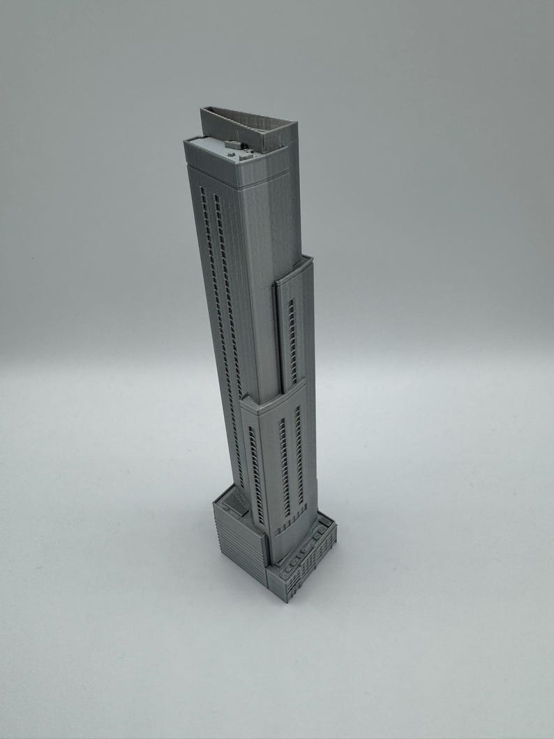 May include: A gray, miniature skyscraper model with a rectangular base and a tall, slender tower. The building has many windows and a flat roof. The model is made of a solid material and is set against a plain background.